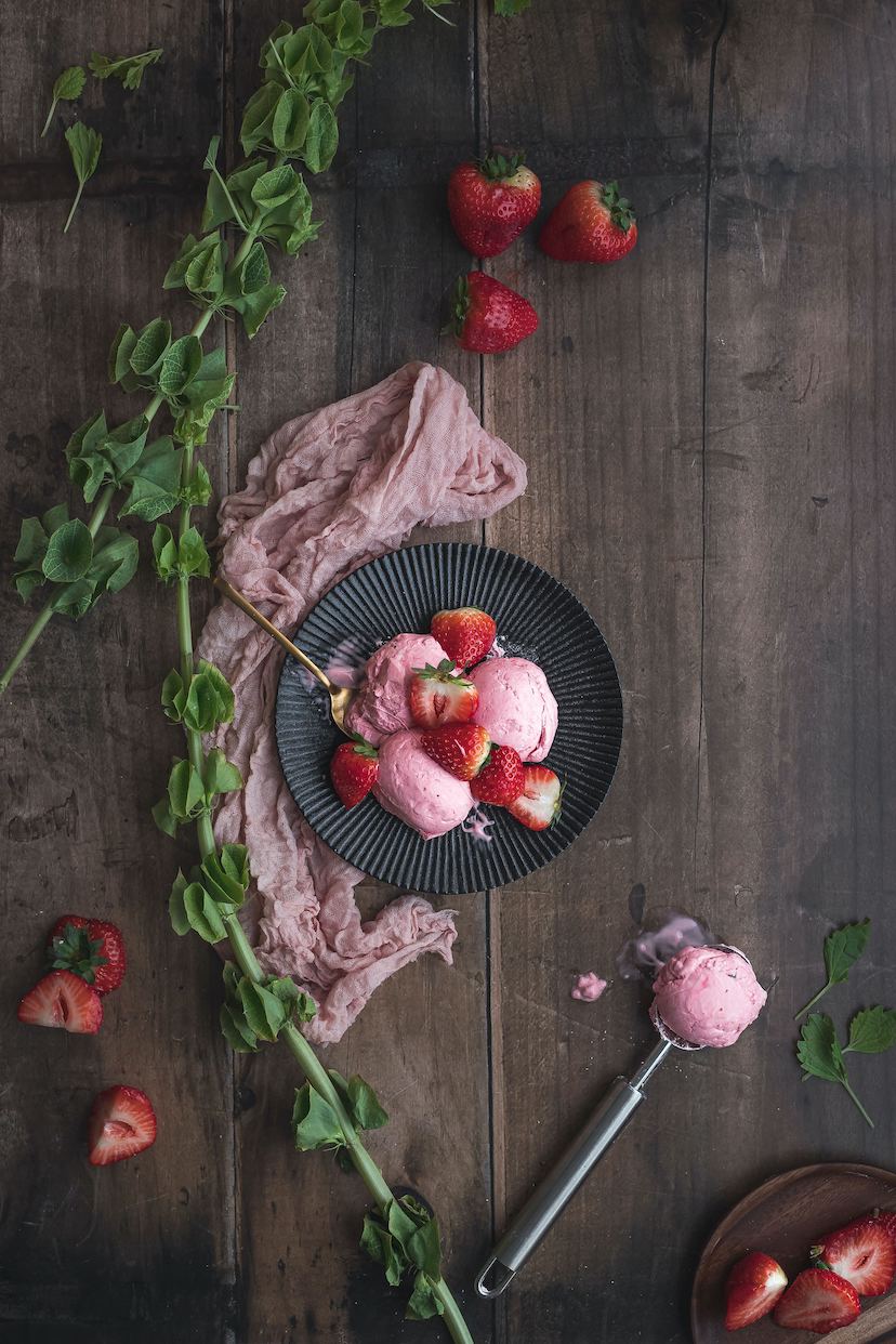 a black plate of pink strawberry ice cream made with Adri Wellness' hibiscus powder placed on a dark wooden surface with fresh strawberries, pink table runner a scoop and fresh plants