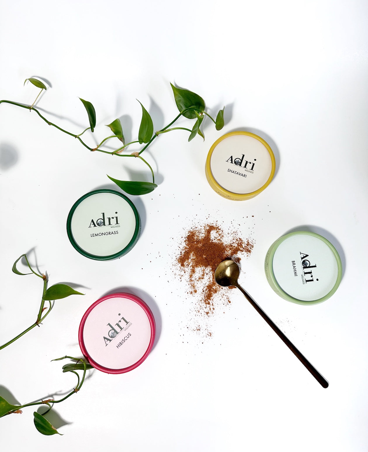 a showcase of top lids of Adri Wellness' Shatavari, Brahmi, Lemongrass and Hibiscus Powder with some raw hibiscus powder scattered in the middle and some plant leaves showing on the left side of the picture