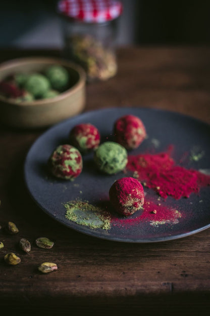 Energy balls made with pistachio powder and Adri Wellness' Beetroot powder placed on a black plate with scattered beetroot powder