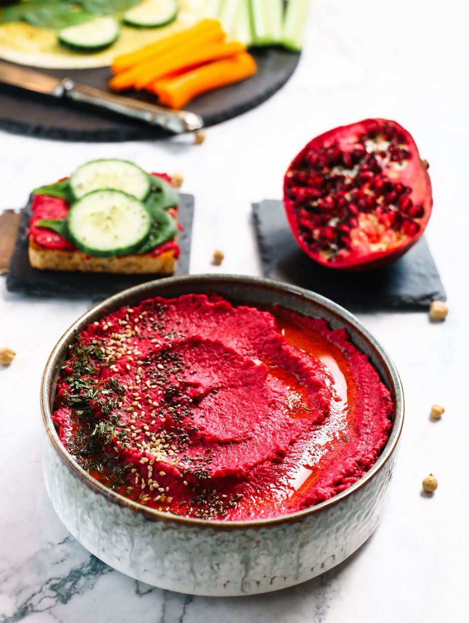 a big bowl of pinkish red hummus made from Adri Wellness' Beetroot powder garnished with chia seeds and sesame seeds placed on a marble surface including a blurry half cut pomegranate in the background with a fresh beetroot hummus cucumber toast
