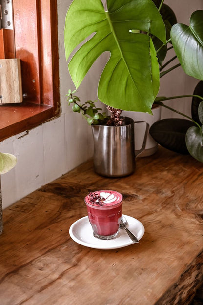 a small transparent glass of pink latte made with Adri Wellness' Beetroot powder including white latte art placed on a white plate with a steel spoon on a kitchen counter