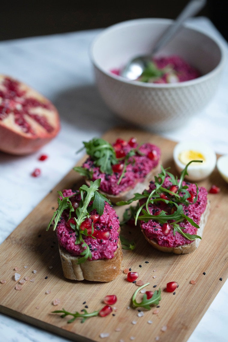 fresh pieces of bread with a spread made from Adri Wellness' Beetroot powder placed on a wooden serving board topped with fresh arugula, pomegranate and hard boiled eggs