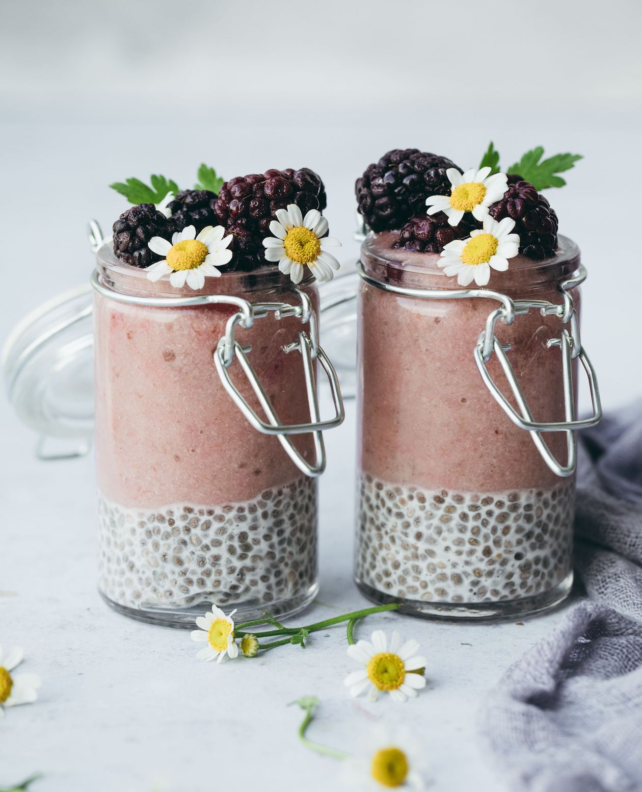 two tall glass jars of chocolate chia pudding made with Adri Wellness' chamomile powder garnished with fresh blackberries and fresh chamomile flowers placed on a white surface