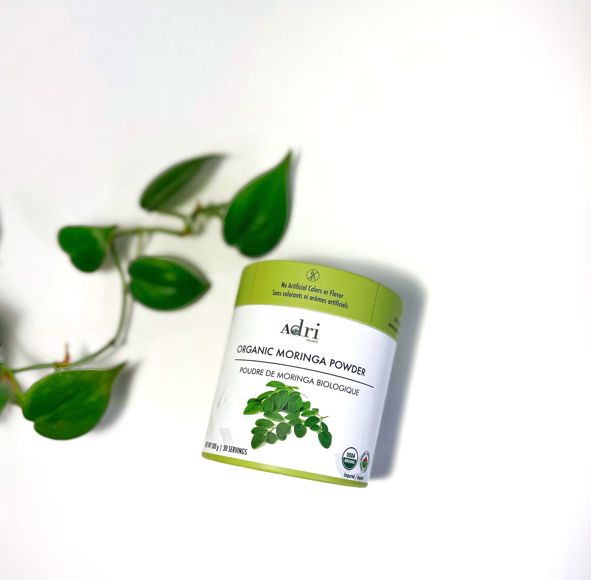 a 100 gm packaging of Adri Wellness' Organic Moringa Powder and fresh plant leaves