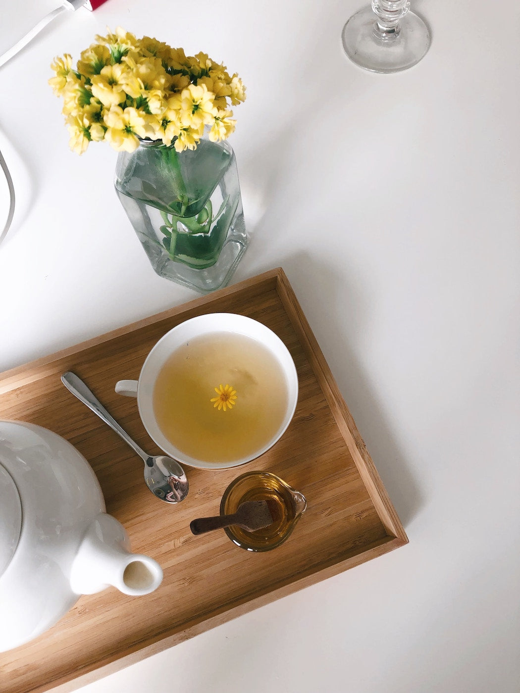 a warm white cup of tea made with Adri Wellness' chamomile powder garnished with a fresh chamomile flower placed on a wooden tray with a white kettle and a spoon