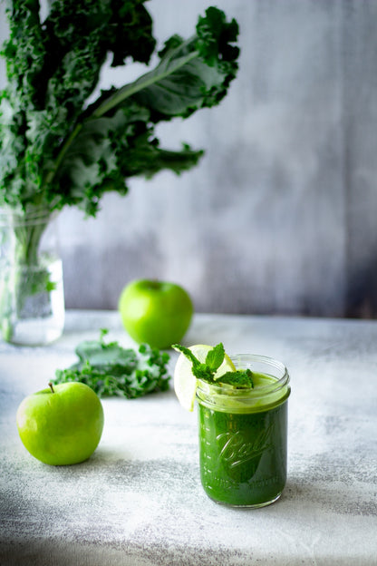 a small glass mason jar of green juice made from green apple, kale, fresh mint, and Adri Wellness' wheatgrass powder placed on a white and grey surface