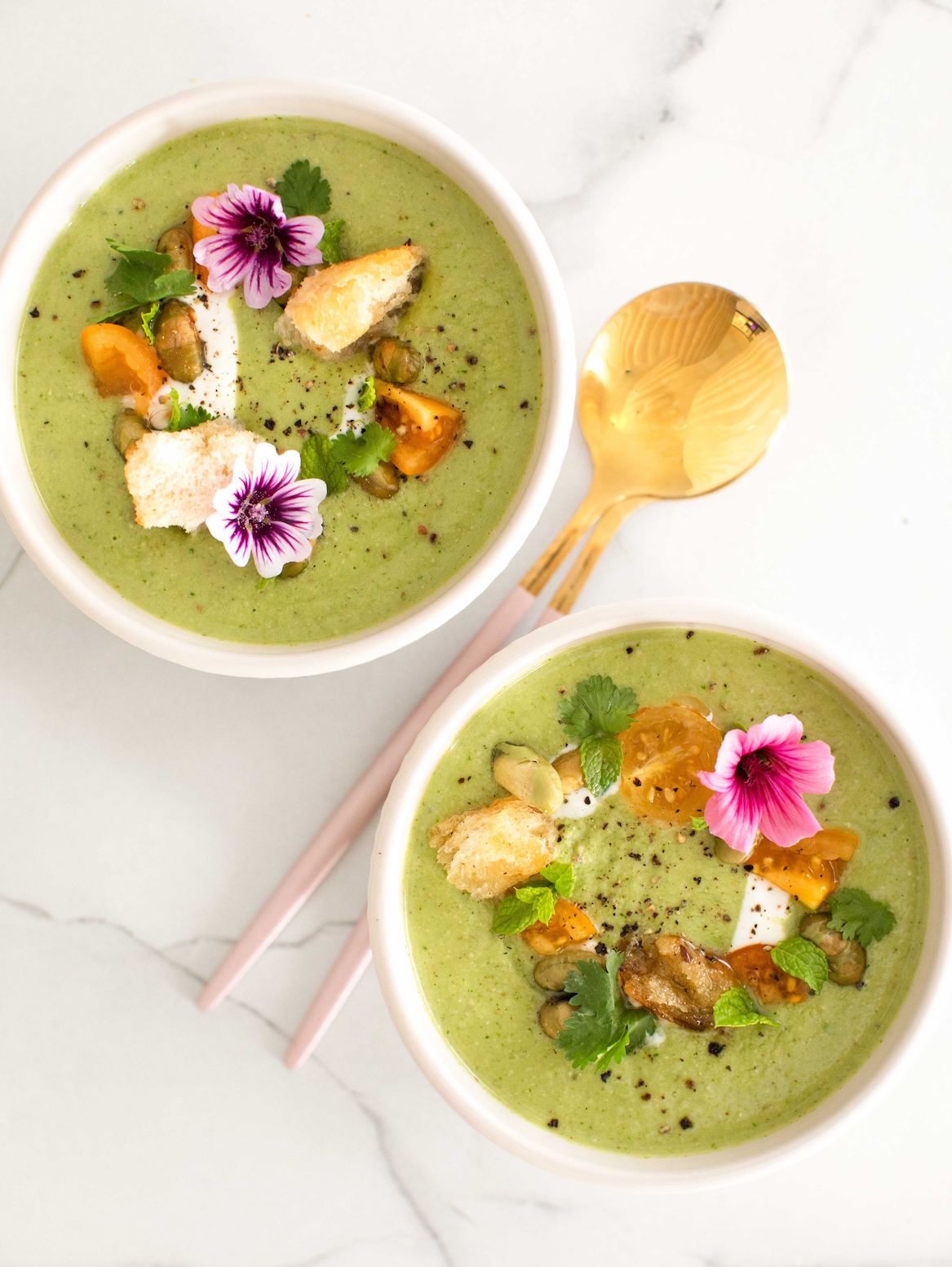 green spinach and pea soup made with Adri Wellness' Lemongrass powder served in a white bowl with a garnish of fresh pink flowers and other herbs placed on a white marble surface with two golden spoons