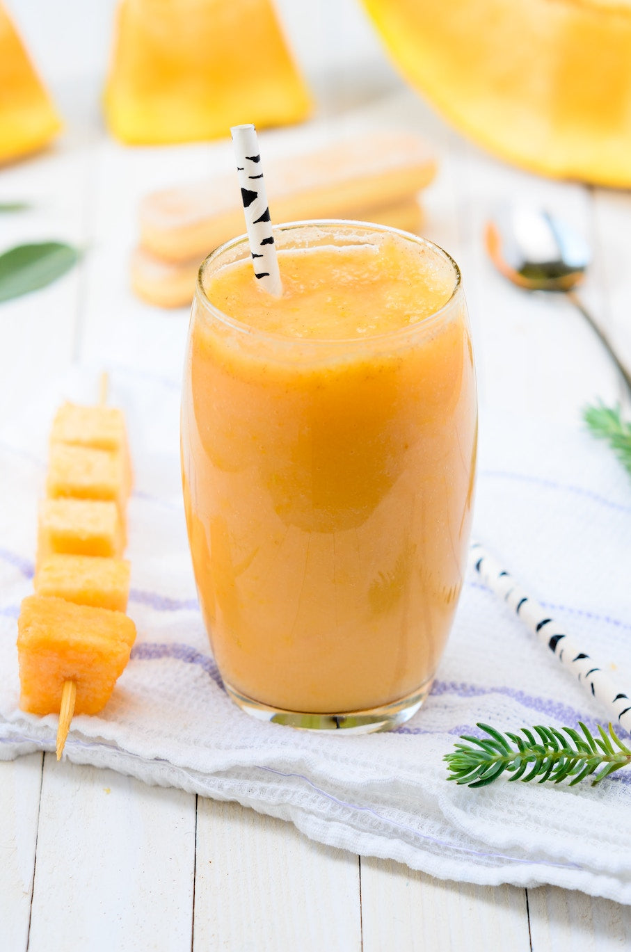 a tall glass of mango lassi served with a black and white straw, made with Adri Wellness' licorice powder including a side of raw mango on a stick